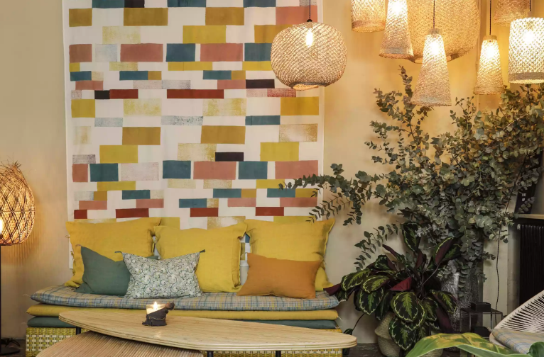 Khu vực tiếp khách với những chiếc gối bằng vải lanh và tấm thảm treo tường bằng vải nỉ màu vàng, xanh lá cây mềm mại. Bộ đèn thả tỏa sáng ấm áp được làm từ vật liệu tự nhiên tạo bầu không khí thư giãn.